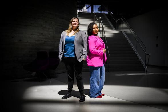 Nita Akoh wearing a pink blazer and blue pants and Jay Nakhai wearing a grey blazer and black pants standing back to back under a spotlight-type light in a darkened room.