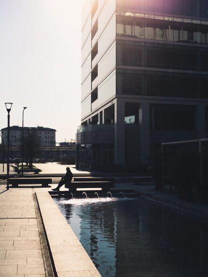 The silhouette of a person sitting on a bench is visible against harsh sunlight coming from the top of a glass office building. The building casts a reflection on a water fountain.