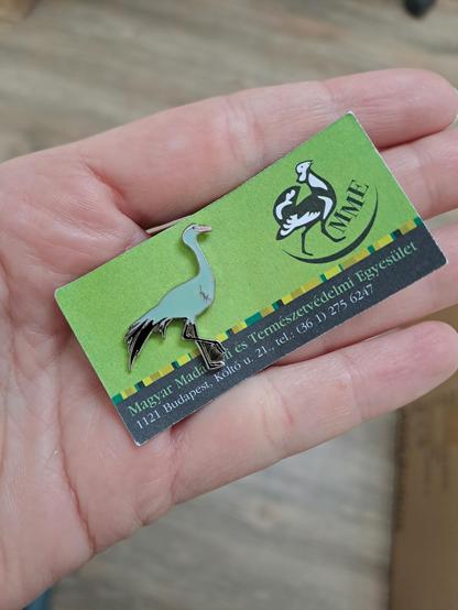 Enamel pin depicting a blue crane, pinned to a green piece of paper with the Hungarian Ornithological Society logo on it.
