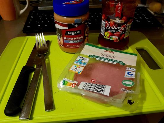 Creamy peanut butter, strawberry preserves, salami,  and cutlery, sit on a green cutting board.