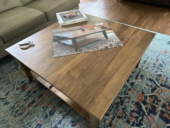 Coffee table with a wooden surface, a remote control, and a completed jigsaw puzzle picturing the same coffee table. The table is positioned on a colorful patterned rug, and a sofa is partially visible in the background.