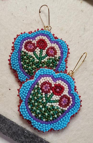 a pair of beaded earrings by kiamichi of flowers with a light blue border, a purple outline, and red and pink stamens, filled with many colors of green and cream colored beads.