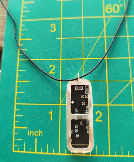 the back side of a silver pendant with an old PCB. it has a black cord and it's placed in green mat