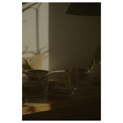 Photo eines gedeckten Küchentisches in der Abendsonne. Eine schwarze Industrielampe hängt über einem alten Kieferntisch. Leere Teller, Besteck ind Gläser zeugen von Personen, die auf dem Bild nicht zu sehen sind. Der Tisch liegt fast vollständig im Schatten, während ein schmaler Streifen Sonne auf der dahinter liegenden Wand die einzige Lichtquelle im Bild ist.