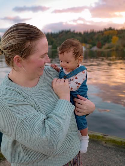 A white non-binary person is holding a small child, smiling at them. The background features a lake with a colorful sunset and trees lining the shore.