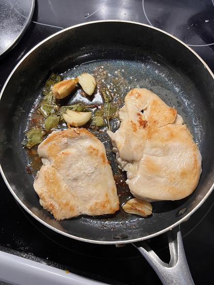 Photo of two chicken breasts cooking in a pan with garlic cloves and fresh sage.