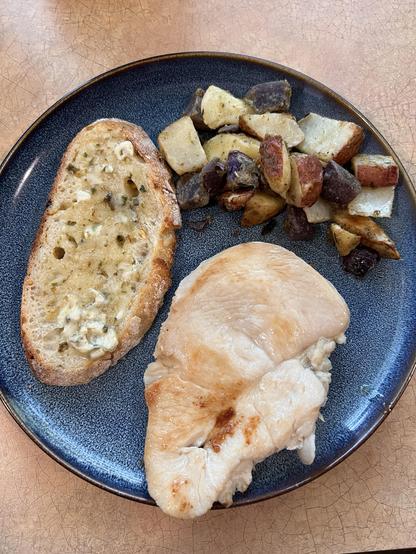 Photo of one of the chicken breasts on a blue plate with some diced and roasted potatoes and a slice of bread topped with an herb butter.
