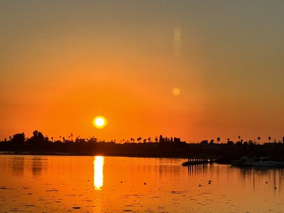 A serene sunset over a calm body of water with the sun appearing as a bright yellow orb low on the horizon. The sky displays a beautiful gradient from deep blue-gray at the top transitioning to vibrant orange and yellow near the horizon. The sun's reflection creates a golden column of light on the water's surface. A dark silhouette of land with scattered palm trees and vegetation spans across the horizon, and there appears to be a small dock or pier structure visible on the right side of the shoreline.