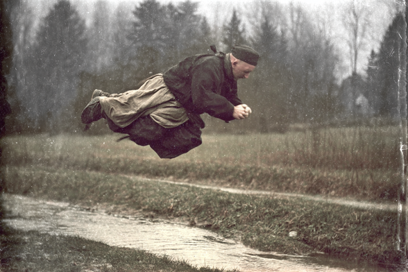 Person horizontally suspended in mid-air over a stream, wearing traditional clothing in a vintage setting.