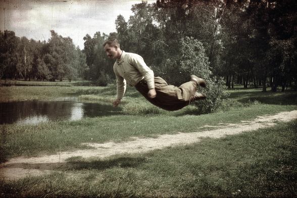 A man appears to float horizontally above a pathway in a green park, with trees and a small pond in the background.