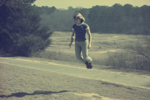 A young person appears to be floating in mid-air above grass with a path and trees in the background.