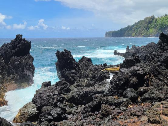 Seashore with jagged, black volcanic rock formations in the foreground, turquoise and blue ocean behind. In the far background, a point covered in green vegetation juts out into the ocean.