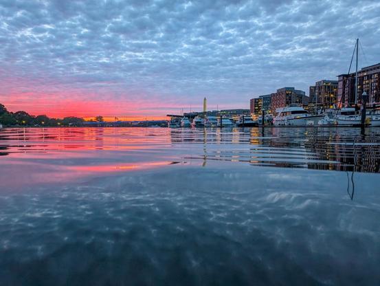 Pink horizon at dusk and numerous low clouds in light blue sky reflect in calm water with gentle waves and the Washington monument in the background and the wharf buildings to the right of frame.