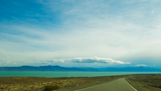 a wide, dramatic landscape is photographed. a road stretches to a vanishing point in the lower right and beyond, a turquoise lake is seen stretching across the horizon in front of a lineof blue mountains. above, some clouds are seen in a hazy-white sky with some blue patches
