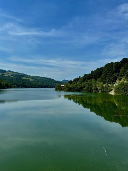A tranquil lake surrounded by green hills under a clear blue sky. The water reflects the landscape, creating a serene atmosphere.