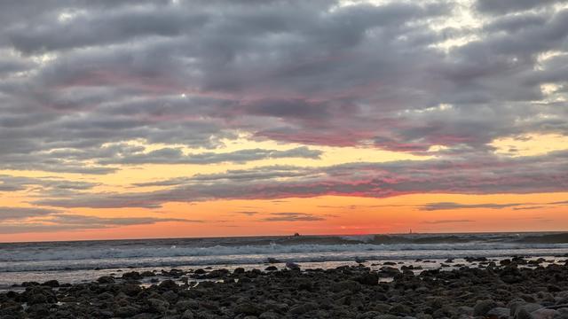Historical Documentation in the form of a photograph of a rising sun over the ocean bay at Yirrell Beach in Winthrop, MA. The sky is filled with vibrant colors of orange, pink, and purple, with the sun rising in the distant horizon. The clouds are scattered across the sky, creating a dramatic and moody atmosphere. The ocean is calm and the waves are crashing onto the shore. In the foreground, there is a rocky beach with small pebbles scattered across it. The overall mood of the image is electric and awe inspiring an amazing start to the new day.