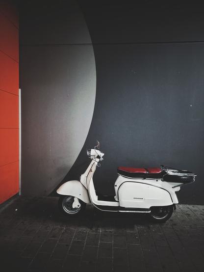 The portrait-orientated image shows a white scooter with a red seat parked on a paved surface. The paving is made of dark square tiles. Behind the scooter is a dark gray wall with a white semi-circle. To the left of the dark gray wall is a vertical orange surface.