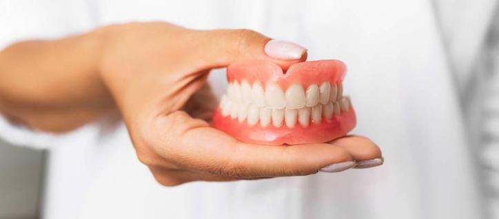 A close-up shows a woman holding a full set of dentures in the palm of her hand. The dentures have white teeth and a pink gumline. The woman is wearing a white lab coat, and her fingernails are painted in a subtle iridescent color.
Photo from free stock images.