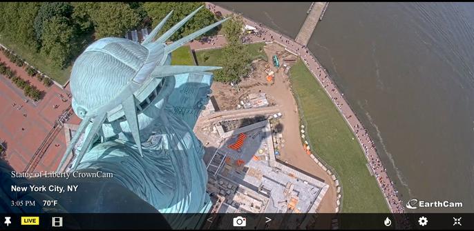 View from the torch of the Statue of Liberty today. The crown of the “Statue of Liberty Enlightening the World” is lit by the sun, while below, construction materials appear at the base of the statue and visitors can be seen on the walkway. Trees and grasses on either side of the image are deep green in color. The Statue of Liberty is made of copper, which has oxidized, turning from brown to a light green.