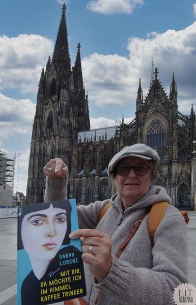 Frau zeigt auf ein Buch, das sie hält. Im Hintergrund eine Kathedrale. Das Buch heisst: Mit dir möchte ich im Himmel Kaffee trinken. Auf dem Cover ist ein Frauengesicht. Die Autorin des Buches ist zu lesen: Sarah Lorenz.