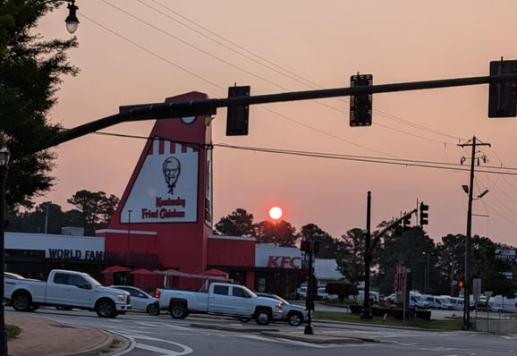 A morning sun in orange color against haze from Canadian Wildfire smoke at a busy intersection with cars at the bottom, a big Kentucky Fried Chicken sign on a huge chicken landmark.