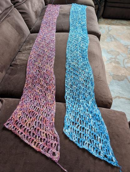 two partially-finished crochet wraps, one in variegated orange/salmon/purple and the other in variegated blue/teal/white, on a brown couch