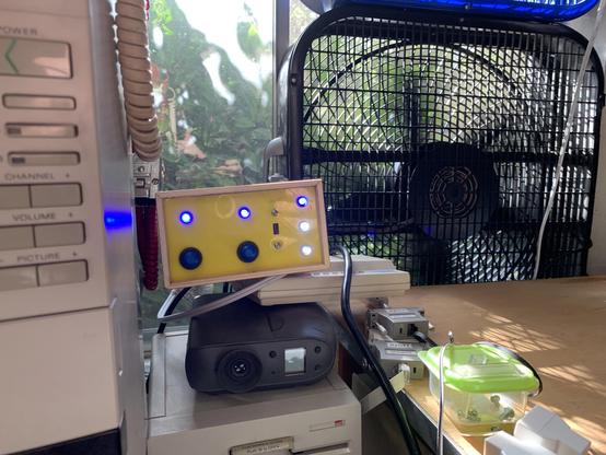 Outlet controller with yellow face plate, blue & white LEDs, and blue buttons, with fans in background. Blue LEDs are on indicating everything is powered up and fans are running.