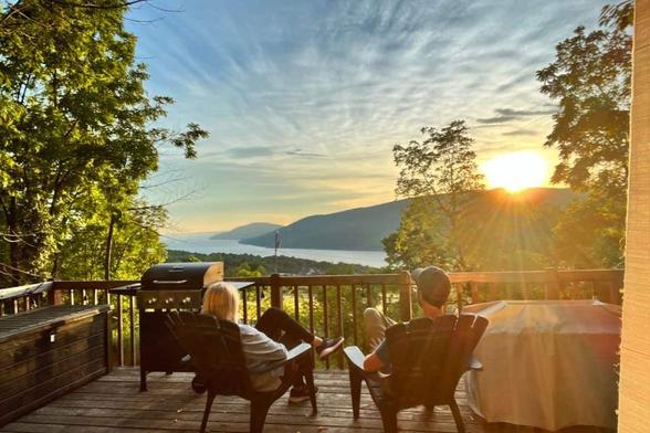 A man and woman sit on a deck, watching the sun rise over rolling hills along a peaceful lake in the Finger Lakes, NY.