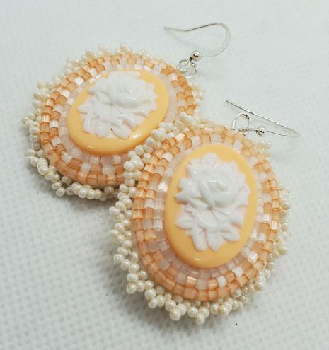 an elegant pair of beaded earrings. the centerpiece is a cream colored rose on a pastel orange background. its surrounded by clear, shimmery pale orange, and pearlescent beads to accentuate it