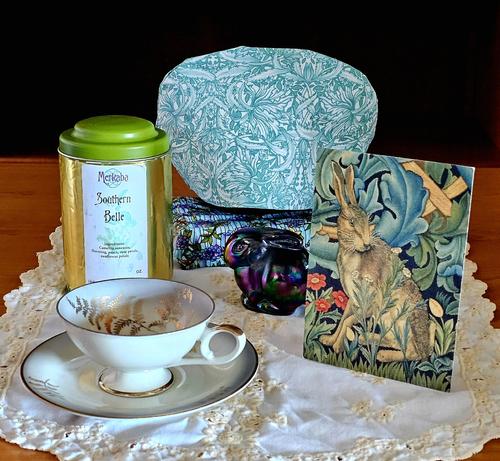 Tea tableau on ivory runner with eyelet lace upon which are a Fenton amethyst carnival glass bunny paperweight, tapestry style artwork of a hare in a garden patch, elegant antique cup and saucer, and William Morris style tea cozy on a Tiffany stand. The tea is a green and black loose-leaf blend flavored with peach and marigold from Merkaba in Idyllwild, California