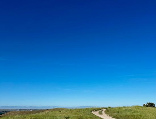A winding dirt path leads through a grassy hillside under a clear blue sky. The landscape is open and serene, with distant mountains visible on the horizon.