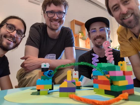 Four people sitting around a table, facing the camera. In front of them is a little construction made out of colorful toy bricks, connected with funny fluffy wires which symbolize peer-to-peer connections