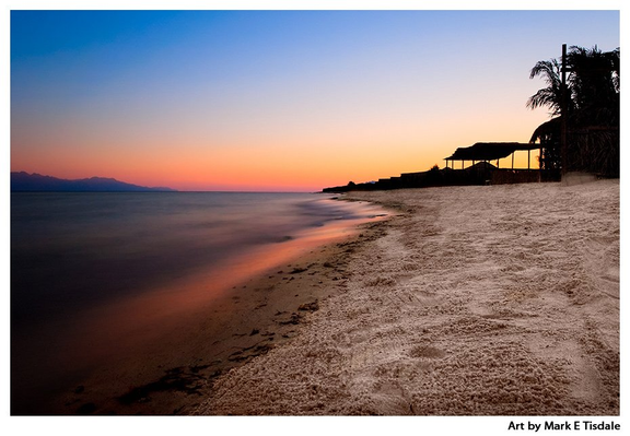 Sunset scene with water in one half of the image and a sandy beach in the other. A small beach hut and a palm tree are seen in silhouette. the sky is pink and blue.