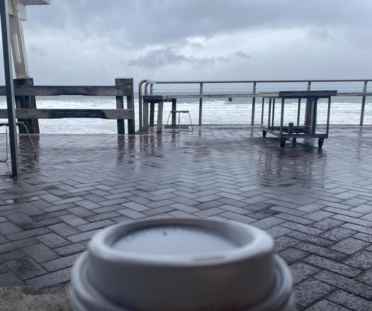 Top of a takeaway coffee in foreground with a view across the wet terrace to a cold and grey ocean and sky.