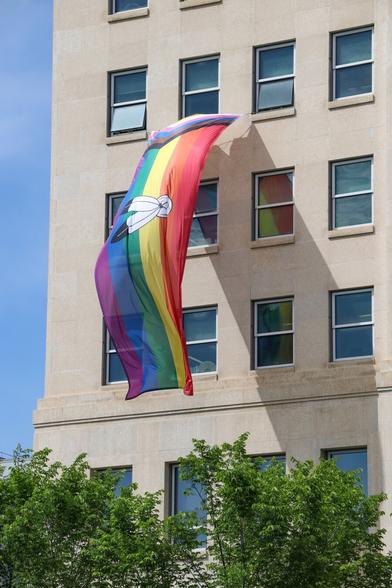 Pride flag with original rainbow stripes, as well as the chevron of stripes representing trans people (white, light pink, light blue) and people of colour (brown, black); in the middle is a black and white image of two feathers representing 2-spirit Indigenous individuals.