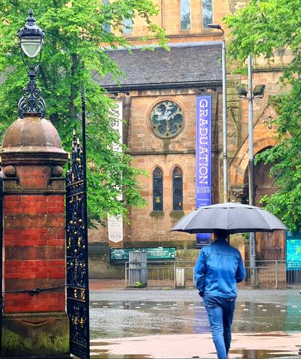 A person with an umbrella walking through a set of Victorian gates towards a traditional sandstone church in the west end of Glasgow