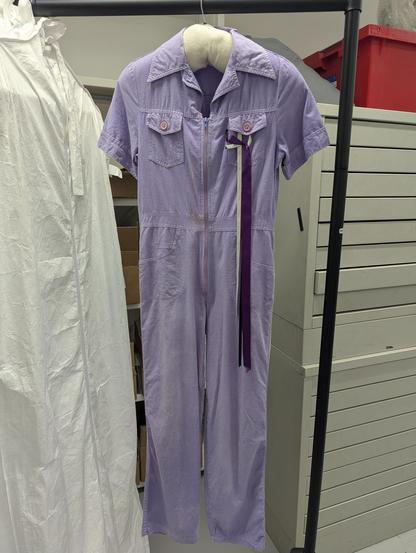 A photograph of a purple jumpsuit hanging over a rail in a museum objects store. You can see other clothing on the rack covered in protective white sheets, and a draw of banners in the background.  The jumpsuit is a faded purple colour, with some ribbons hanging off one of the pockets. There are patches of mud on it left over from the march that it was worn for.