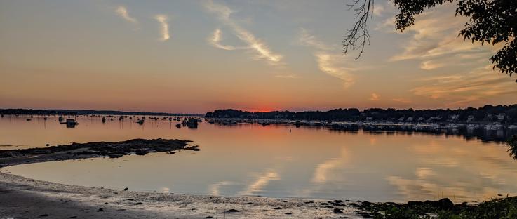 Historical Documentation in the form of a photograph of a rising sun over the ocean bay at the Historic Pioneer Village in Forest River Park, Salem, MA. The sky is filled with vibrant orange and yellow hues, with the sun rising in the yonder. The water is calm and reflects the colors of the sky, creating a mirror-like effect. On the right side of the image, there is a rocky shoreline with trees and bushes on the left side. In the distance, there are several boats docked in the harbor. The overall mood of the photograph is a bit odd but cool.