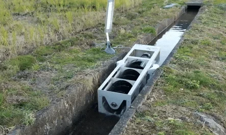 A white-framed PicoPica water turbine generator positioned in a shallow rocky stream, showing multiple dark spiral turbine blades inside the rectangular housing designed to capture energy from slow-moving water flow.