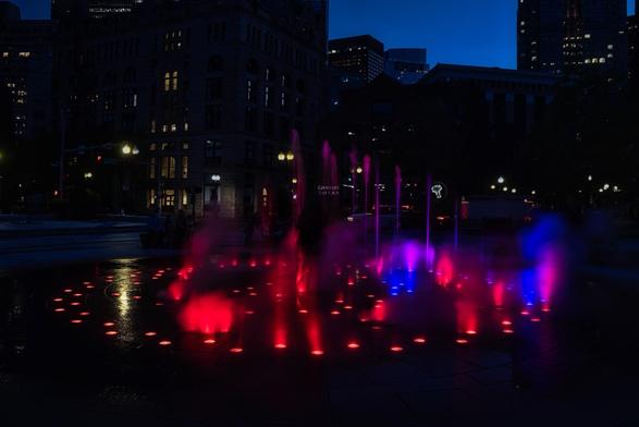 A vibrant water fountain illuminated with red, blue, and purple lights creates a colorful display against a darkening cityscape. Nearby, people are seated, and buildings are partially visible, adding to the urban ambiance.