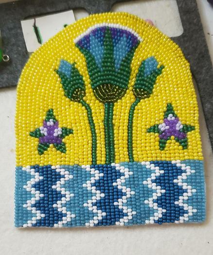 a mostly completed in-progress beading project by kiamichi of an Egyptian design made with seed beads. the color scheme is teal, blue, white, green, and purple with yellow in the background. it depicts an Egyptian lotus coming out of the Nile with some decorative blooming stars next to it