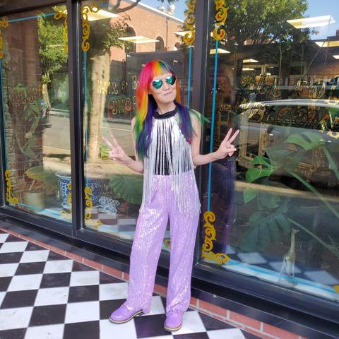 A person(me) with rainbow colored hair and heart-shaped sunglasses stands in front of a store with a window decorated with yellow and red swirls. The person is wearing a black top with a silver fringed vest, sparkling lavender pants, and matching shoes. They are making peace signs with both hands. The ground is made of black and white checkered tiles. Reflected in the window are trees, a brick building and lettering that partially reads “HAIR & SHOP”.