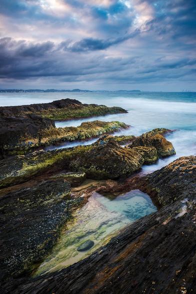 A vertical twilight seascape. Diagonal rock platform and rock protrusions covered in moss. Long exposure smooth water. Distant city and hills in the background under the moody sky.