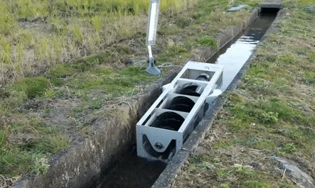 A white-framed PicoPica water turbine generator positioned in a shallow rocky stream, showing multiple dark spiral turbine blades inside the rectangular housing designed to capture energy from slow-moving water flow.