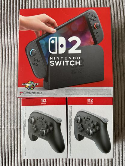 Auf dem Foto ist die Verpackung einer neuen Nintendo Switch 2 zu sehen. Die Konsole wird gerade von einer Hand aus der schwarzen Dockingstation herausgezogen. Das Design ist überwiegend in Rot und Schwarz gehalten. Auf dem Bildschirm der Konsole sind bunte Lichtreflexe sichtbar. Auf der Verpackung steht „Nintendo Switch 2“ sowie ein Hinweis auf das Spiel „Mario Kart World“, das offenbar beiliegt. Unter der Hauptverpackung stehen zwei Originalverpackungen von Nintendo Switch 2 Pro Controllern nebeneinander. Alles liegt auf einem grauen, gerippten Stoff, wahrscheinlich ein Sofa oder eine Decke.