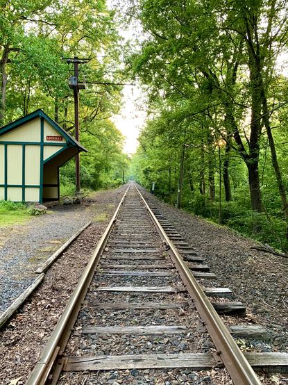 A view of train tracks extending straight into the woods under a partly cloudy sky. A small wooden train station, painted light yellow-beige, stands on the left.