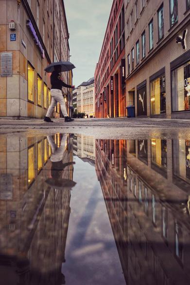 Das Bild zeigt eine städtische Straßenszene an einem bewölkten Tag. Eine große Pfütze im Vordergrund reflektiert die umliegenden Gebäude und eine Person, die mit einem geöffneten schwarzen Regenschirm die Straße überquert. Diese Person trägt eine dunkle Jacke und helle Hosen, was in der Reflexion besonders deutlich wird. Auf der linken Seite befindet sich ein Gebäude mit der Hausnummer 12, das mit mehreren Schildern versehen ist. Auf der rechten Seite sieht man verschiedene Geschäfte, darunter ein Puma-Laden. Die Straße ist von hohen Gebäuden gesäumt, die in die Ferne führen und eine urbane, moderne Atmosphäre schaffen.