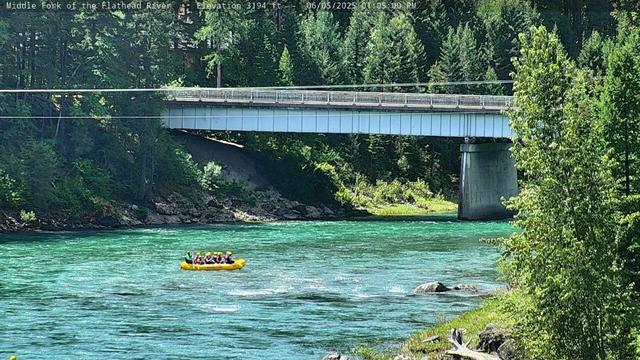 Rafters on the Middle Fork of the Flathead River in Glacier National Park today. The colors of the river include green, white, black and grey. Trees on either bank are free of snow. In the background is the West Glacier Bridge. The raft floating in the river is yellow as are the helmets of the park visitors in the raft.