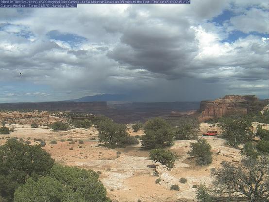 This US Geological Survey research camera in Canyonlands National Park shows a blue sky with light and dark clouds today. Below, park visitors in a red vehicle drive through green trees and vegetation. The exposed geology is tan, brown, grey, and white.
