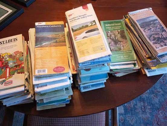 A color landscape photo of five stacks of folded paper maps sitting on a table. The five stacks are different sizes. Stacks one and two are the same height on the left. The map on top of stack one says, "St. Louis," and on stack two says, "Northern California." Stack three is the tallest with forty or more maps. The top map says, "Southern Nevada and Death Valley Area." Stack four to the right of three is the shortest and it says, "Bridger Teton National Forest." Stack five, on the far right says, "Indian Country," on the top map.
